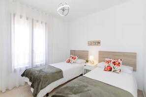 Une chambre d’hôtel avec deux lits, une table de chevet avec une lampe et une fenêtre avec des rideaux légers.