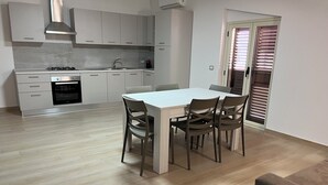 A modern kitchen with white cabinets, a gas stove, and a built-in oven. There is a dining table with four chairs and a window with blinds. - Kalon Eidos. The magical place of Ionian Calabria (San Carlo-Condofuri Marina)