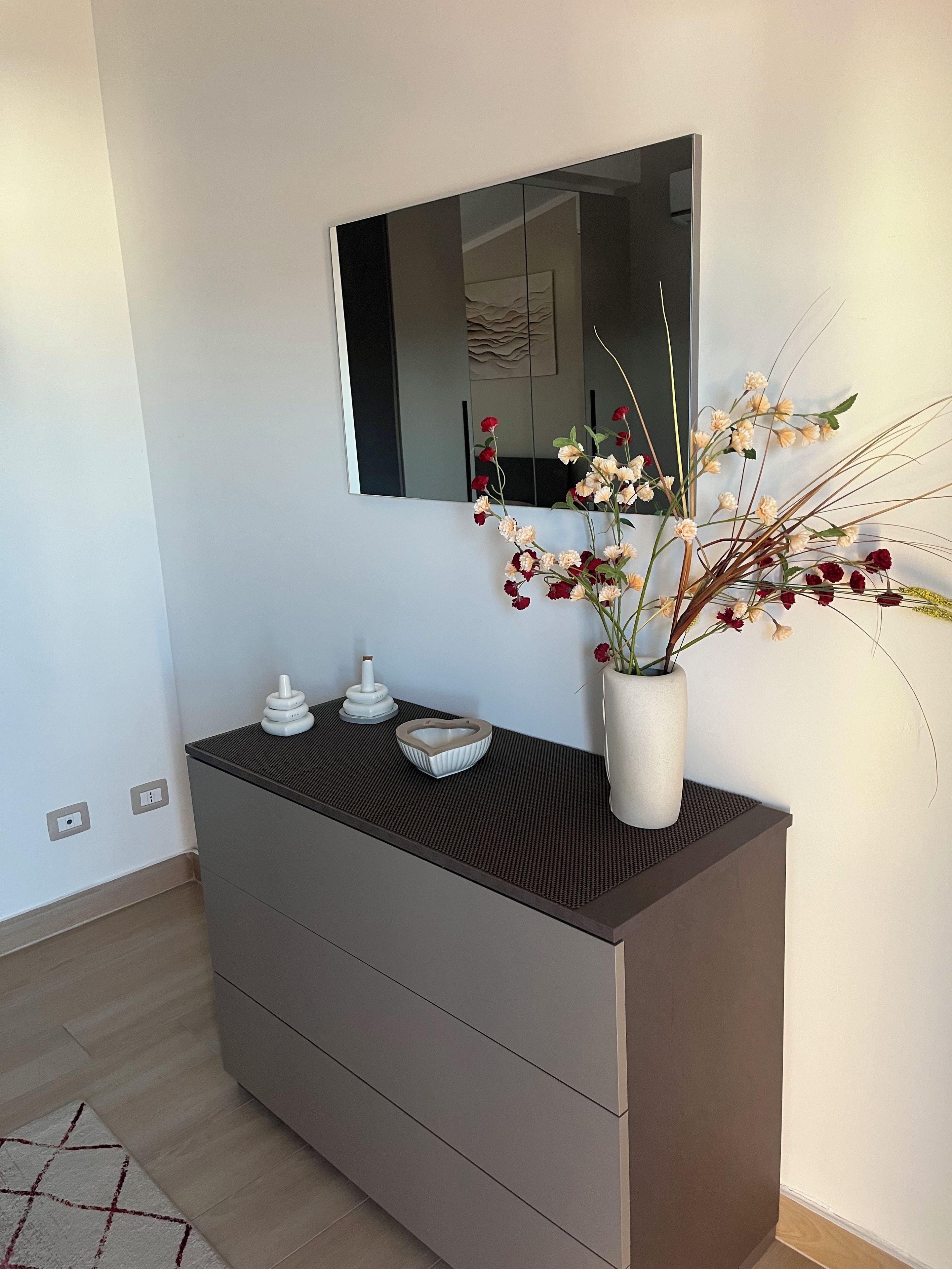 A modern bathroom with a dark-colored vanity, a mirror, and a vase with flowers.