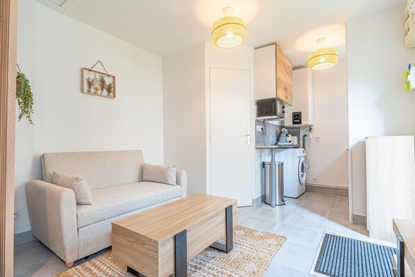 A compact living space with a sofa, a wooden coffee table, a kitchen area with a microwave, a washing machine, and a refrigerator.