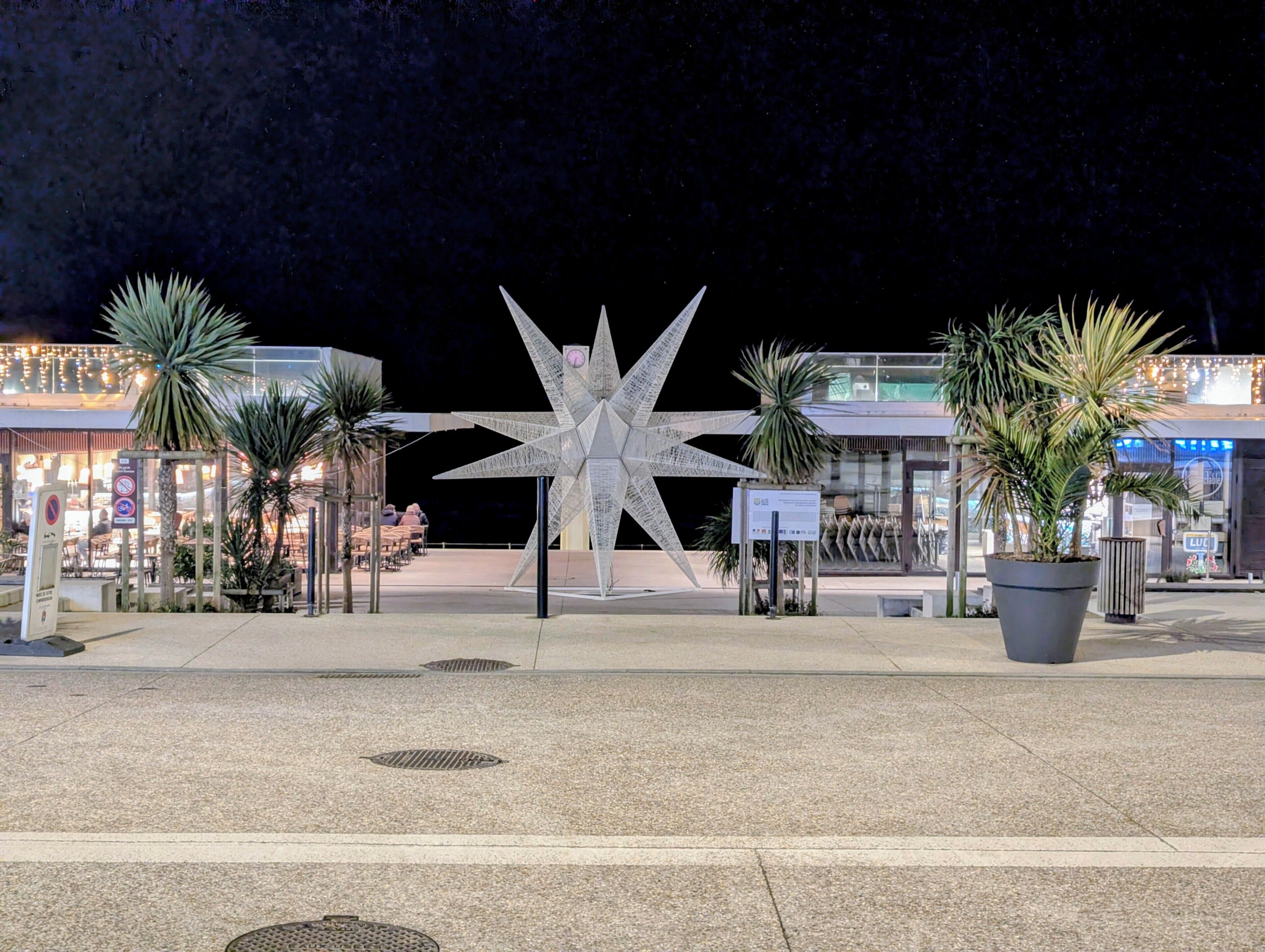 Una escultura con forma de estrella en una plaza, con palmeras y tiendas al fondo.