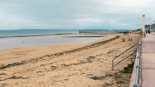 A beach with a pier extending into the sea, a walkway with a railing, and a clock tower.
