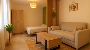 A modern living room with a sofa, a smaller bed, a wooden coffee table, and a hanging plant.