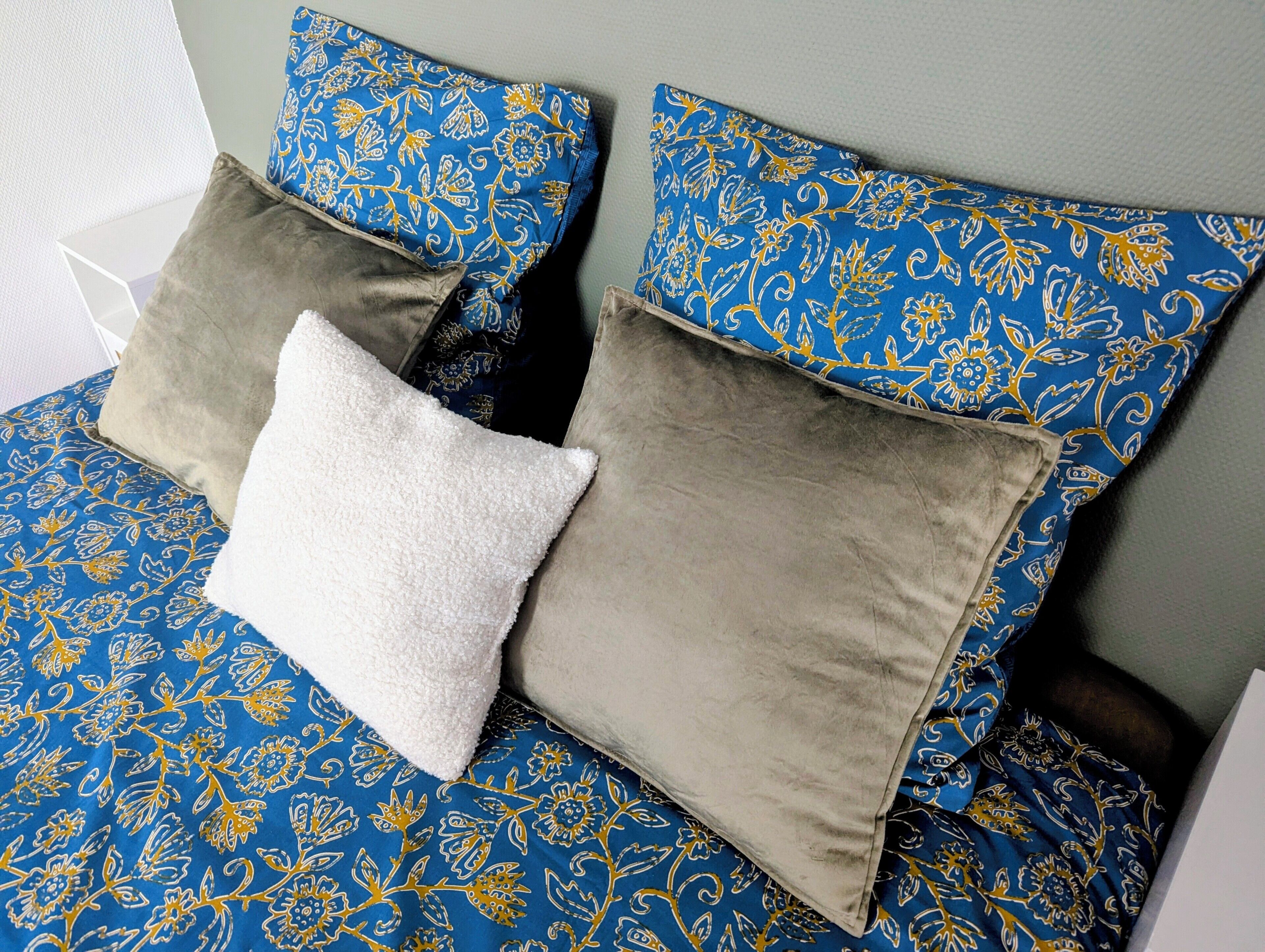 A bed with a blue floral bedspread, two pillows with a blue and gold pattern, and a white pillow.