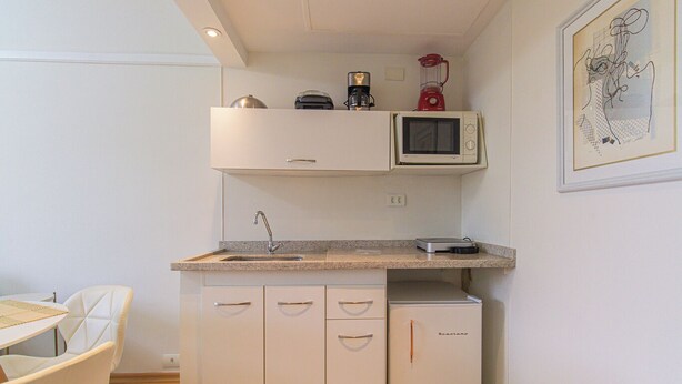 A compact kitchen with a microwave, coffee maker, and blender on the countertop, a small refrigerator, and a dining area with a table and chairs. - Flat c Academia Sauna e Garagem - Jardim Paulista (São Paulo)