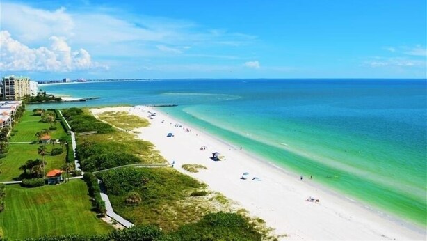 A coastal beach with a clear view of the ocean, a sandy shoreline, and a few buildings in the background. - Beachfront condo at Mansions (TREASURE ISLAND)