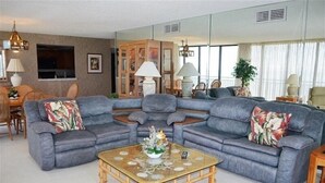 A living room with a grey sectional sofa, a glass coffee table, and a wooden cabinet. - Beachfront condo at Mansions (TREASURE ISLAND)