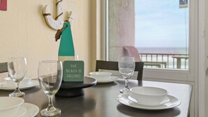 Ett matbord med tomma tallrikar och glas, en skylt med texten ”STRANDEN LOCKAR”, och en utsikt över stranden genom ett fönster.