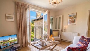A living room with a television, a sofa, a coffee table, and a view of a building through a window. - Chic `Duke` Suite In Historic City Centre (Cambridgeshire)
