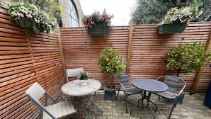 Een terras met een ronde tafel en twee stoelen, omringd door een houten schutting en plantenbakken.