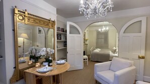 Een kamer met een eettafel gedekt voor twee personen, een witte fauteuil, een kroonluchter en een grote spiegel met decoratieve details.