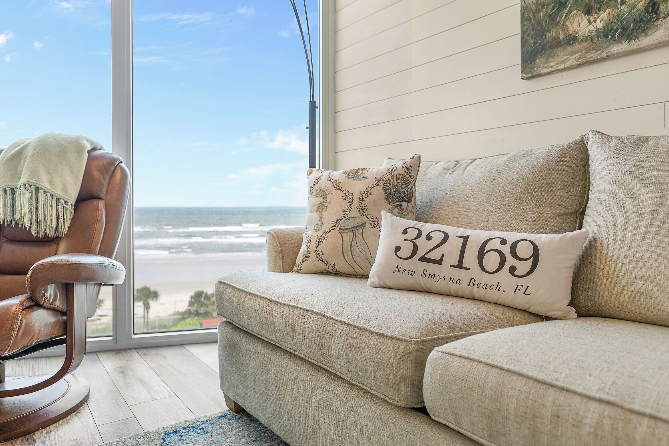 Ein Wohnzimmer mit Blick auf den Strand, ein Ledersessel und eine Couch mit dekorativen Kissen.