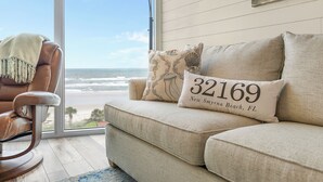 A beige sofa with a cushion that reads "32169 New Smyrna Beach, FL" and a view of the ocean through a large window. - Golden Arms 602 (New Smyrna Beach)