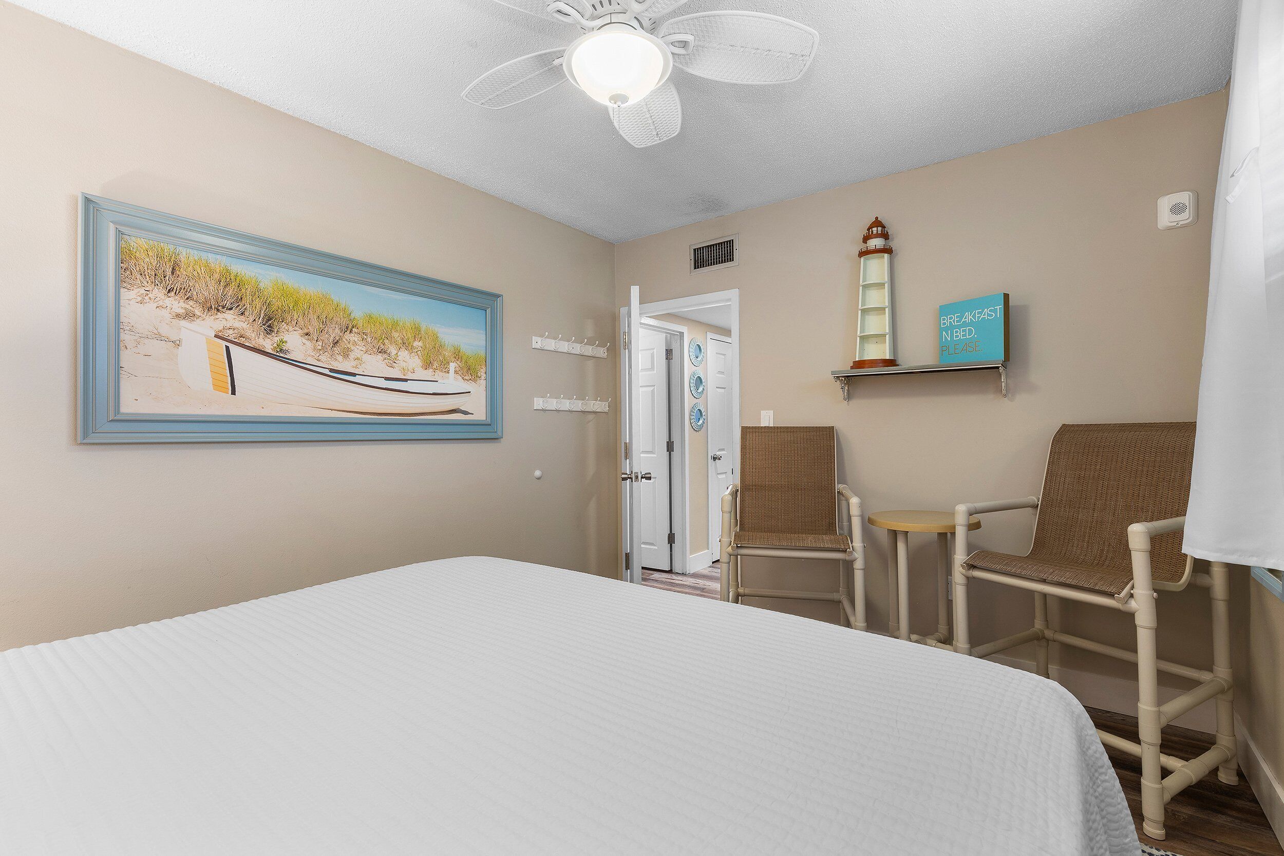A hotel room with a bed, two wicker chairs, a painting of a beach scene, a ceiling fan, and a sign indicating breakfast is served.