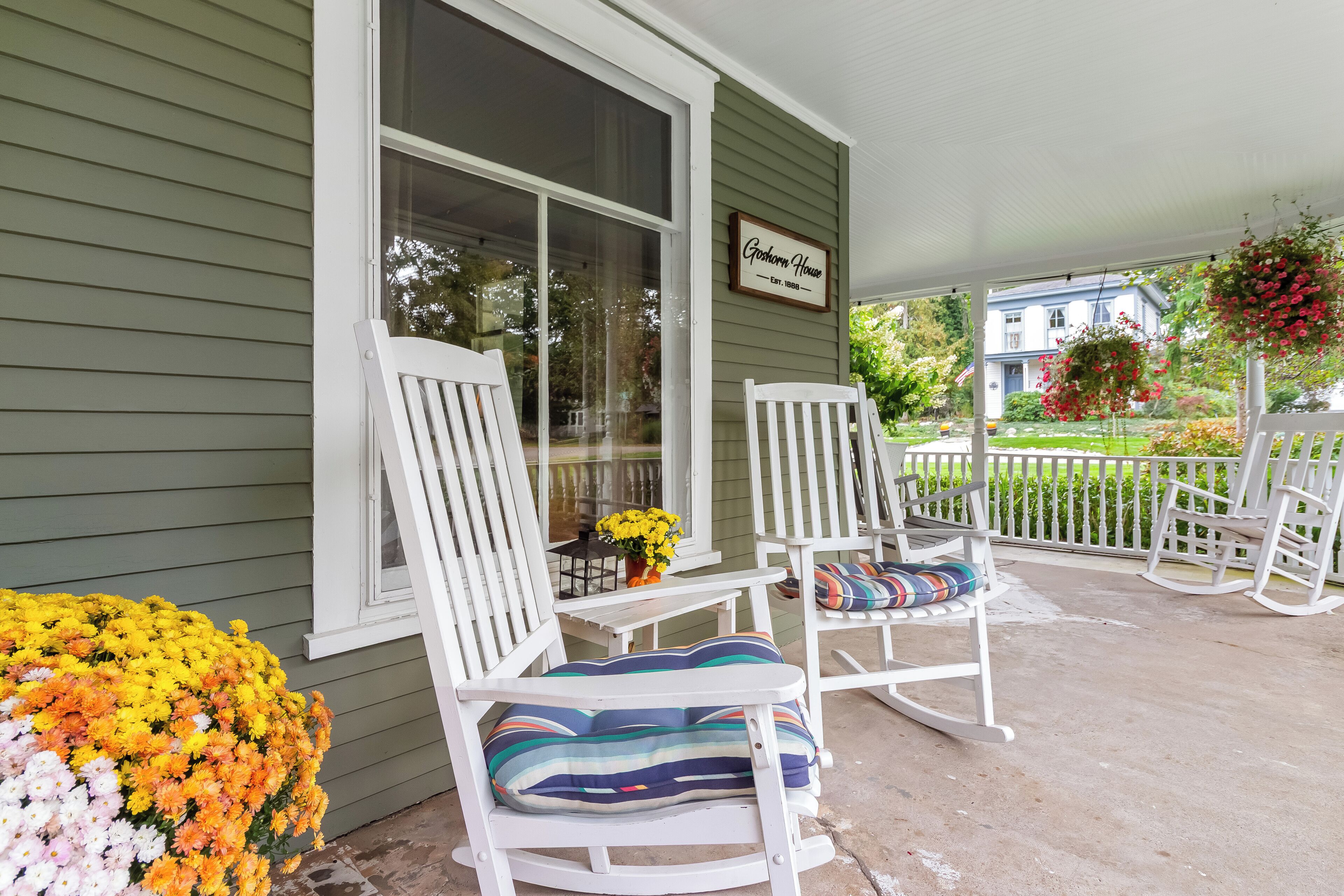 A porch with two rocking chairs, a small table with a vase of flowers, and a sign that reads 'Goshen House.'