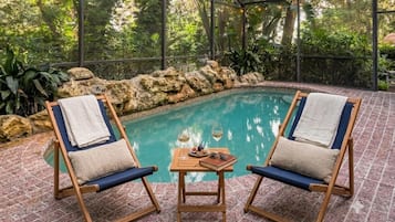 An indoor pool area with two lounge chairs, a table with glasses and fruits, and a view of lush greenery through large windows.