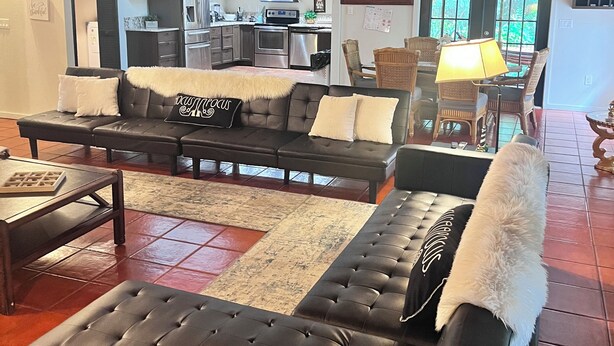 A living room with a sectional sofa, a coffee table, and a rug. There is a kitchen in the background with modern appliances. - Refuge Home, Jacuzzi & Heated Pool 35 min Orlando. (Eustis)