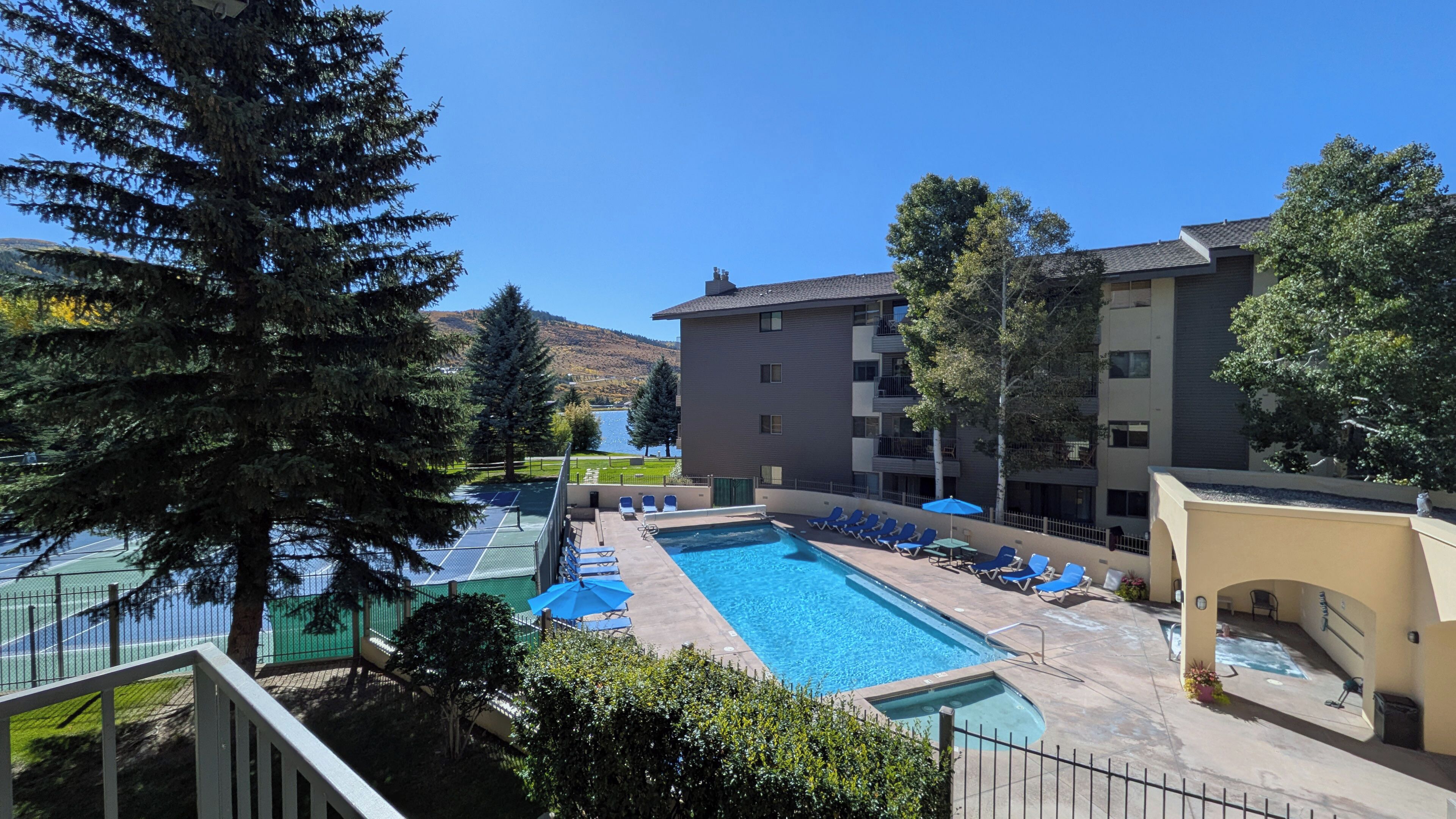 Une piscine avec des chaises longues et un court de tennis, le tout entouré d’arbres et bordé d’un immeuble doté de balcons.
