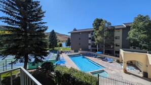 A swimming pool with lounge chairs and a tennis court, surrounded by trees and a building with balconies.