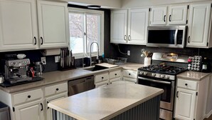 Une cuisine moderne avec des meubles blancs, un îlot central, des appareils électroménagers en acier inoxydable et une fenêtre qui laisse entrer la lumière naturelle.