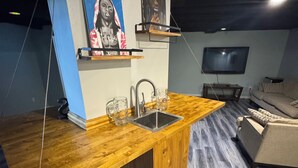 Une cuisine moderne avec un plan de travail en bois, un évier et un meuble intégré. Deux tableaux ornent le mur et un téléviseur est fixé au-dessus d’un canapé.