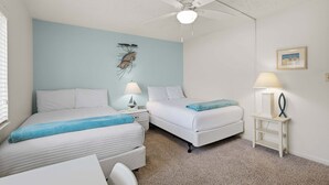 A bedroom with two beds, a ceiling fan, a nightstand with a lamp, and a framed picture on the wall. - Errol By The Sea 126 (New Smyrna Beach)