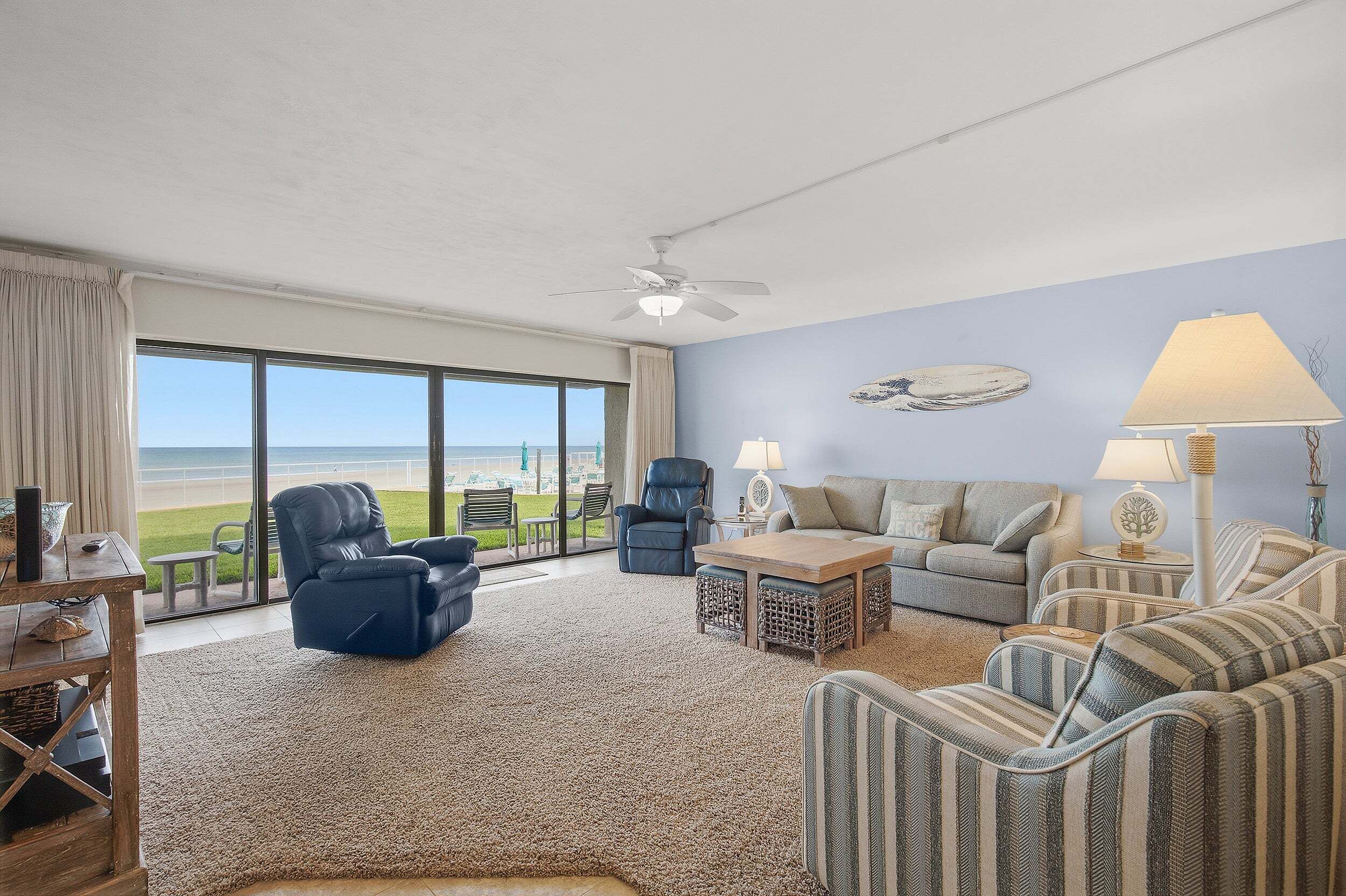 A spacious living room with a view of the beach, a ceiling fan, and a large window.