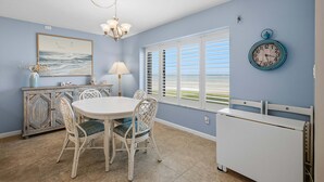 A dining area with a round table and chairs, a chandelier, a painting of seagulls, and a clock on the wall. - Errol By The Sea 126 (New Smyrna Beach)