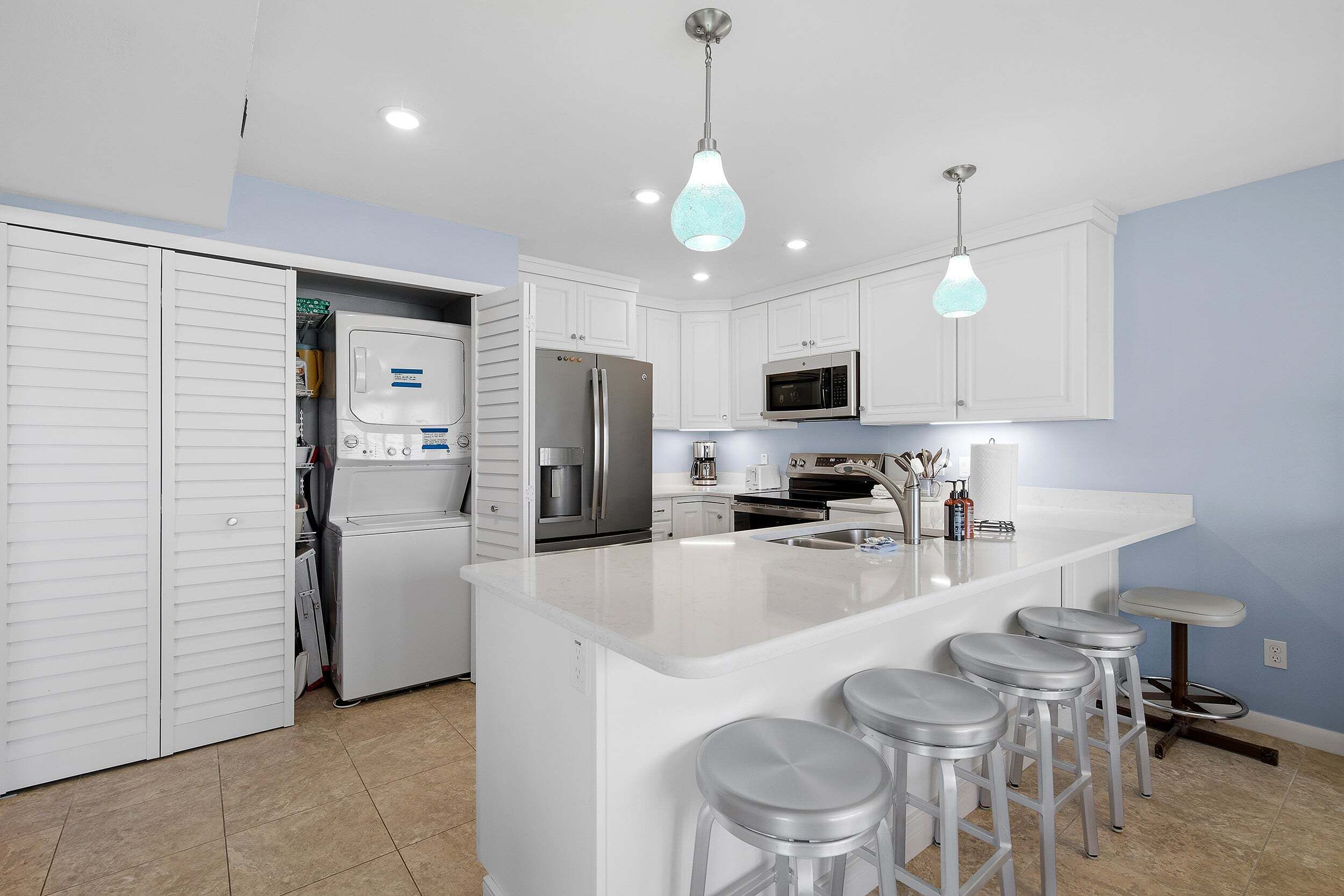 Una cocina moderna con armarios blancos, electrodomésticos de acero inoxidable, una isla central con taburetes y luminarias colgantes.