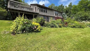 Une maison en bois à deux étages, dotée d’un balcon, entourée d’une végétation luxuriante et d’une pelouse bien entretenue.