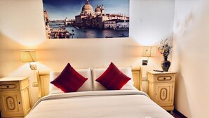 A hotel room with a bed, two red pillows, a nightstand with a lamp, a vase with flowers, and a framed picture of a cityscape. - HOTEL HENRY (Venice)