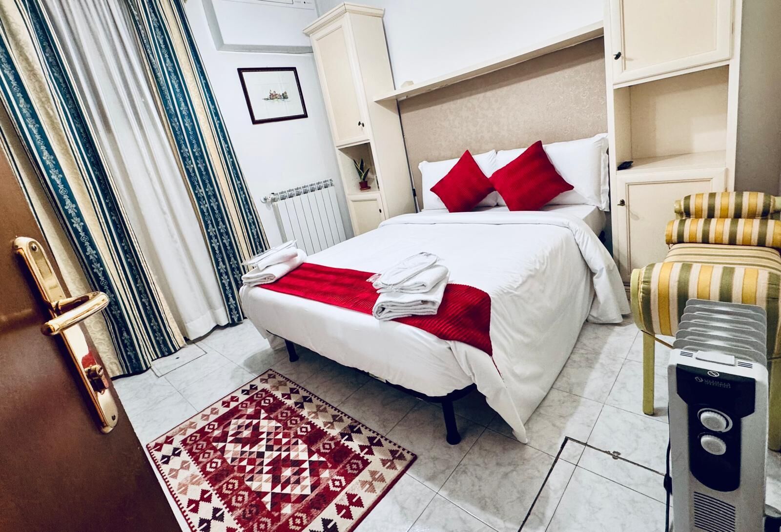 A hotel room with a bed, a red throw blanket, a patterned rug, a radiator, a wardrobe, and a chair.