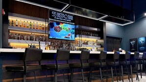 Un espace bar avec de nombreuses bouteilles d’alcool, un écran affichant le menu et des tabourets de bar.