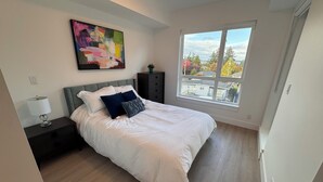 Un dormitorio moderno con una cama grande, una mesita de noche, un aparador, una ventana con vistas a árboles y casas, y un cuadro abstracto de colores en la pared.