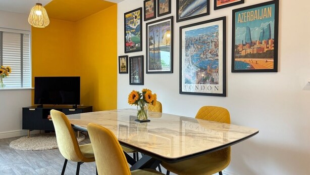 Uma sala de jantar com uma mesa de mármore, cadeiras amarelas, um vaso com girassóis e cartazes de viagens emoldurados na parede.
