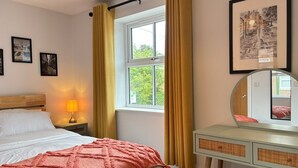 Quarto com cama, luminária de cabeceira, janela com cortinas, cômoda, espelho e quadros nas paredes.