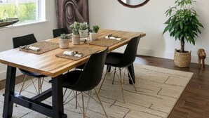 Un coin repas avec une table en bois, des chaises noires, un lustre, un tableau représentant un Bouddha, un miroir rond et des plantes en pot.
