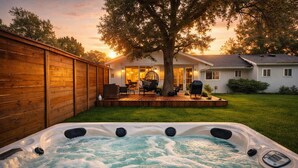 Un jacuzzi dans un jardin, avec une clôture en bois, une terrasse dotée de meubles d’extérieur et une maison en arrière-plan.