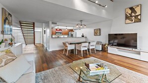 Amplia sala de estar con una mesa de centro de vidrio, un televisor de pantalla plana grande y una zona de comedor con una cocina al fondo.