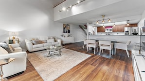 Un amplio salón con un sofá grande, una mesa de centro y un área de comedor con sillas. Se ve una cocina al fondo con electrodomésticos modernos y una escalera que lleva a otro nivel.