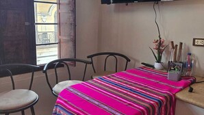 A small room with a wooden table, two chairs, a TV mounted on the wall, and a window with a view of buildings outside. - In the historical center and with all services nearby. (Arequipa)