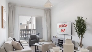 A modern living room with a sofa, a dining table set with plates and glasses, a TV displaying "BOOK ME", and a balcony with a view of buildings. - JVC 1 bed Sleeps 4 With Swimming Pool (Dubai)