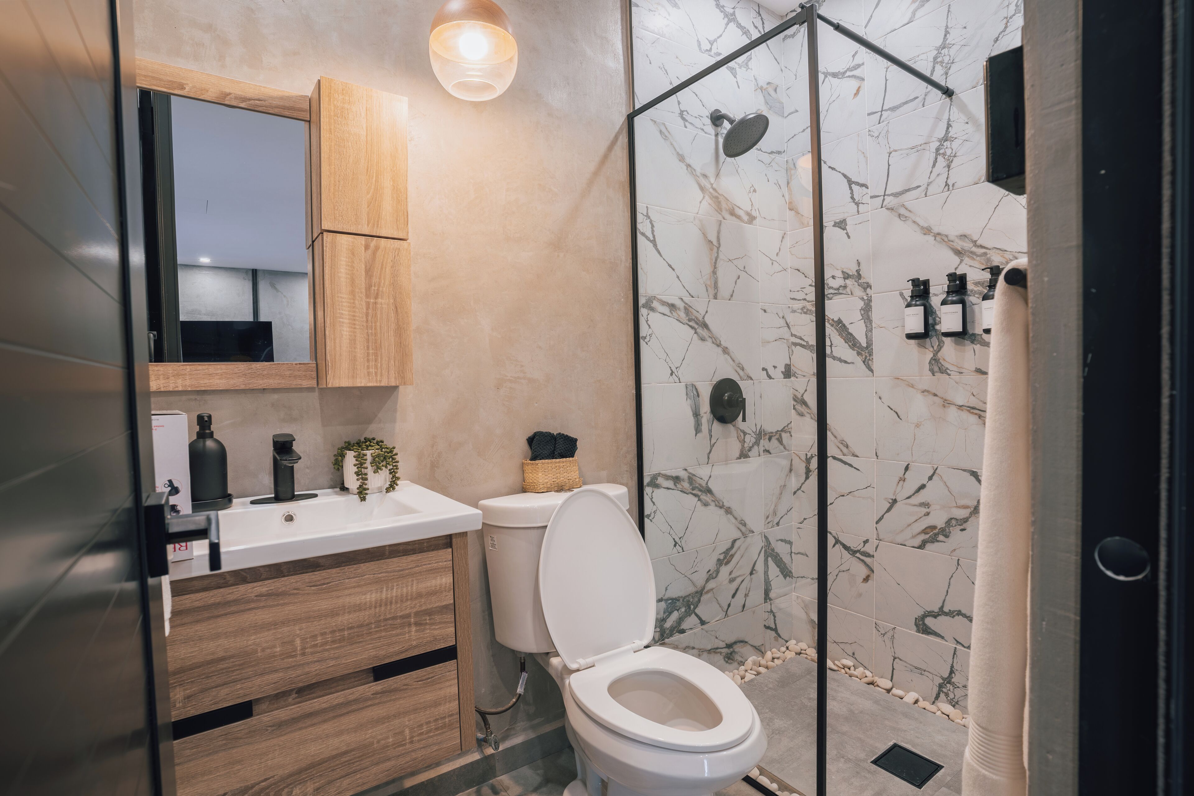 A modern bathroom with a wooden vanity, a white toilet, a glass shower enclosure, and marble tiles.