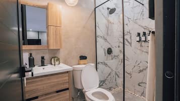 A modern bathroom with a wooden vanity, a white toilet, a glass shower enclosure, and marble tiles.