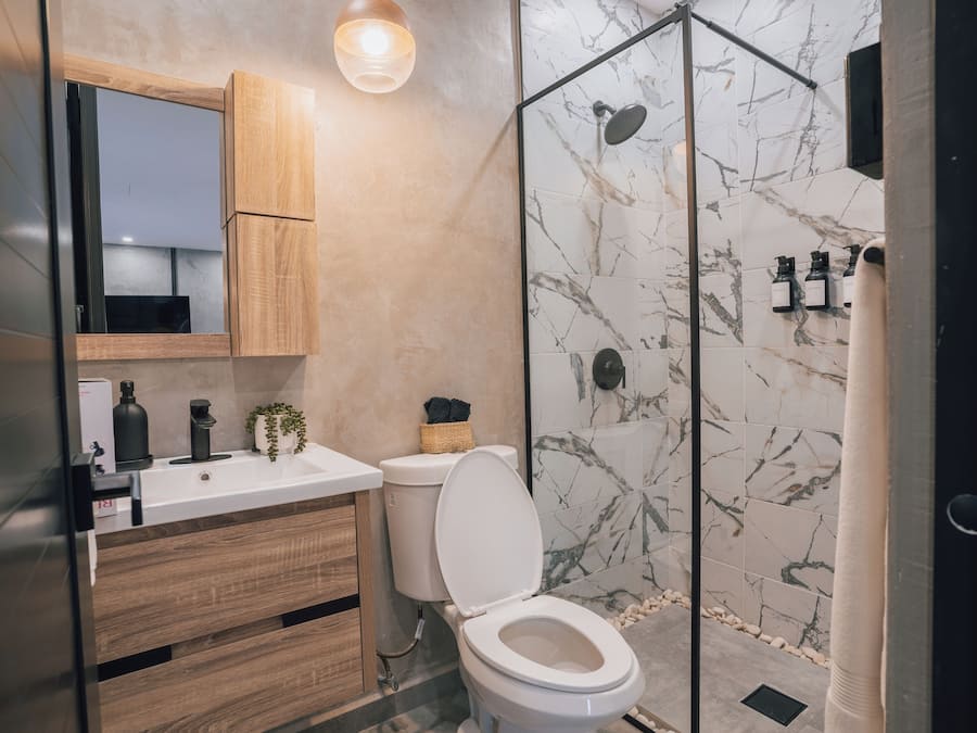 A modern bathroom with a wooden vanity, a white toilet, a glass shower enclosure, and marble tiles.