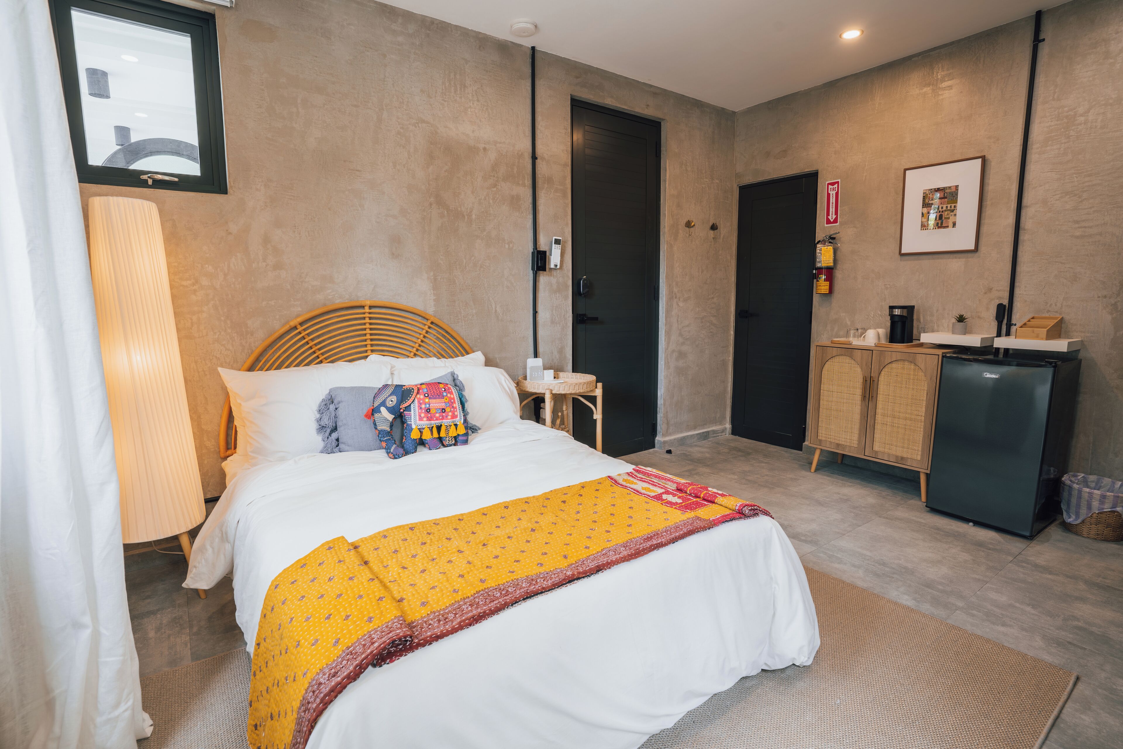 A modern hotel room with a large bed, a bedside table, a wardrobe, and a small kitchenette.