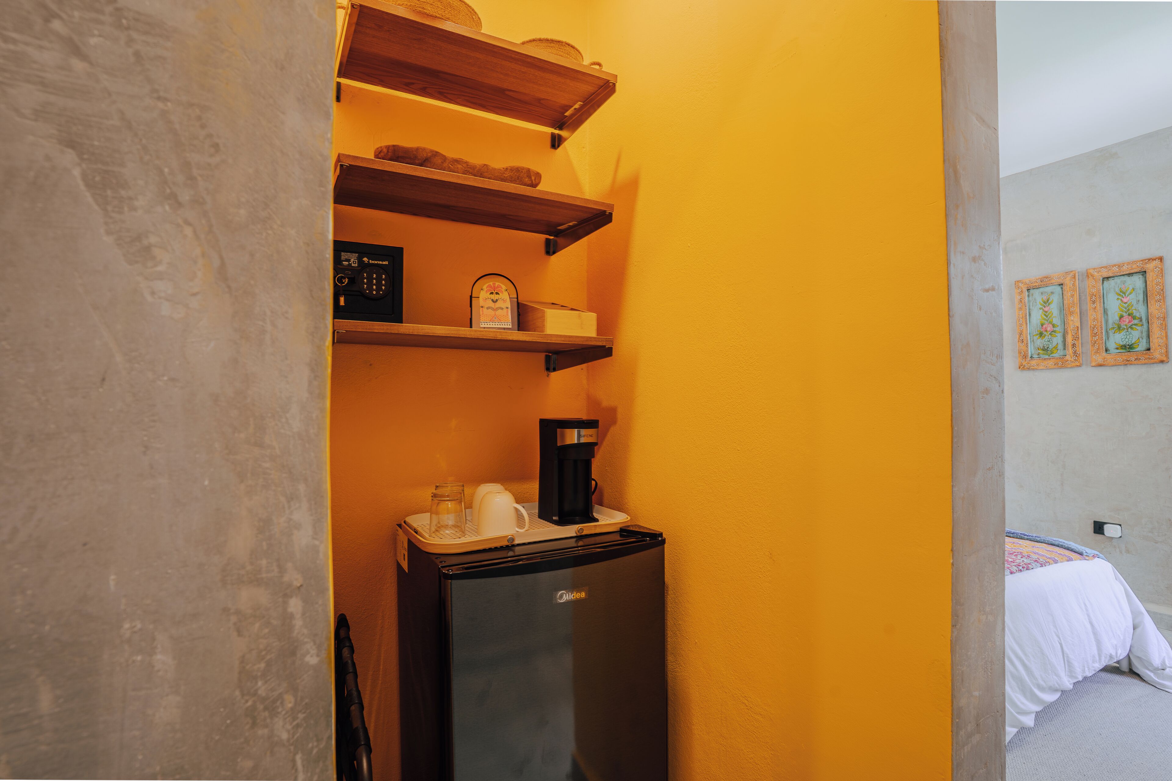 A compact room with a refrigerator, coffee maker, and shelves against a yellow wall.