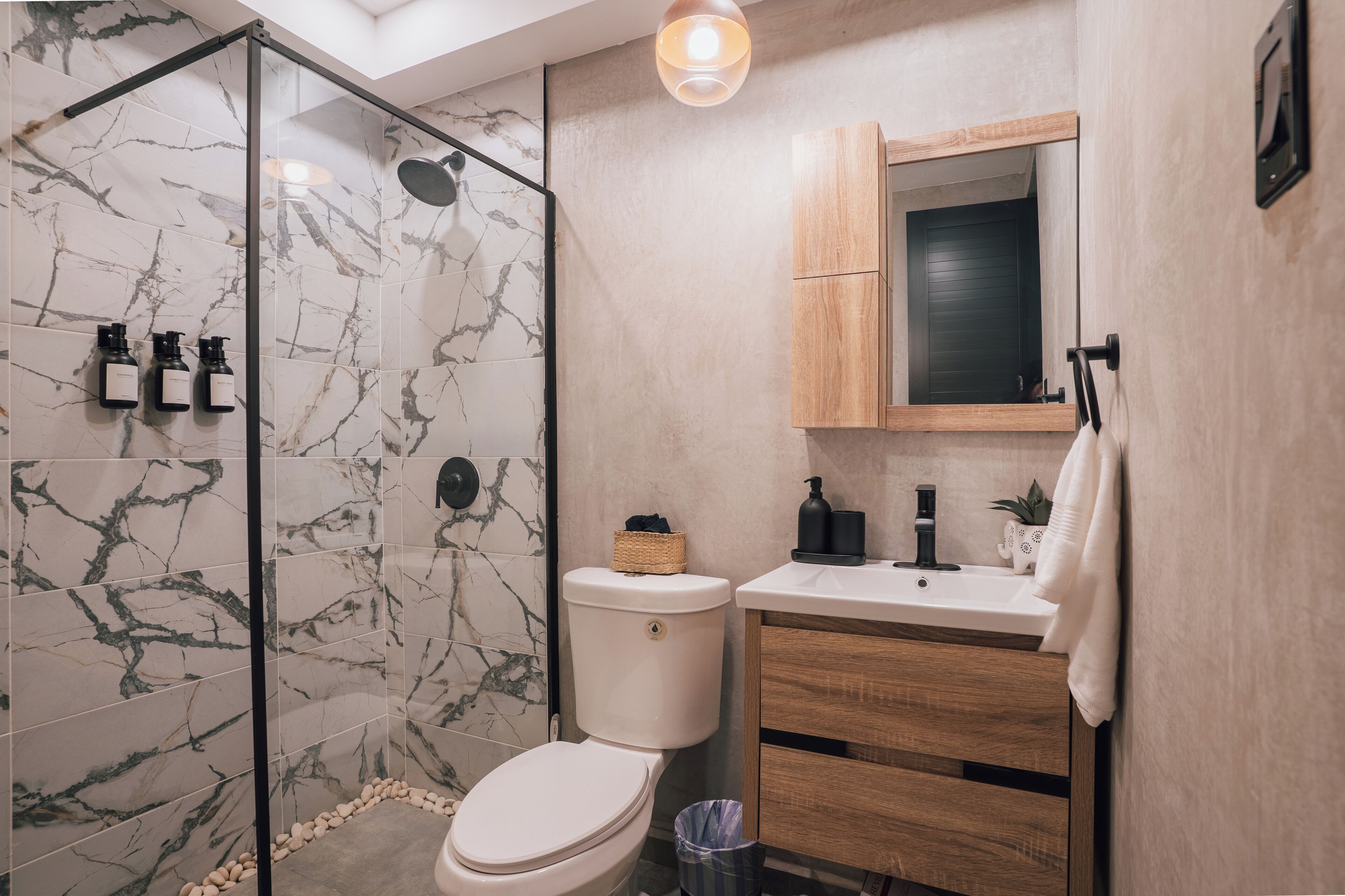 Un baño moderno con ducha envuelta en vidrio, baldosas de mármol, inodoro, lavamanos con mueble de madera y espejo sobre el lavamanos.