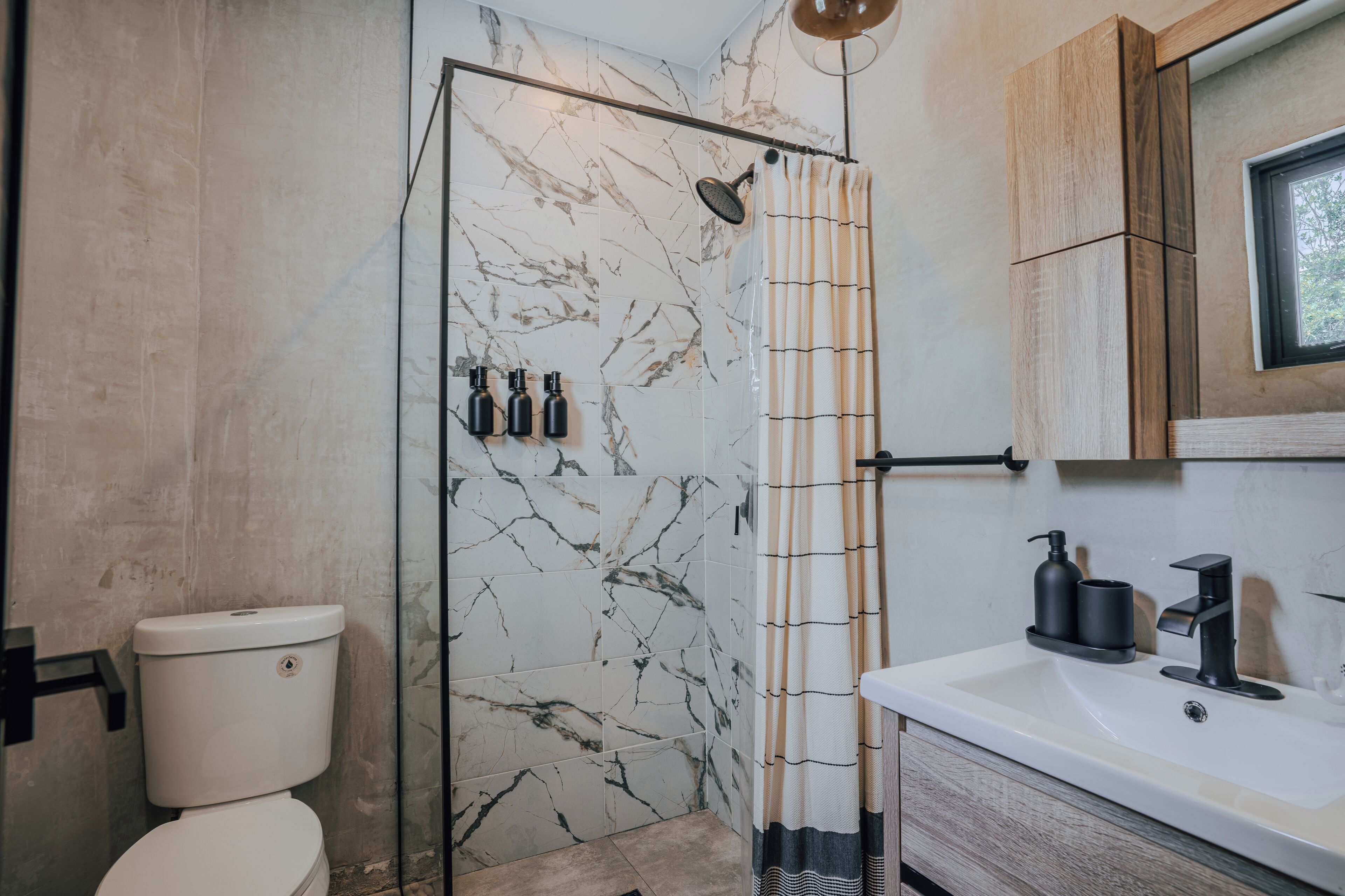 A bathroom with a glass-enclosed shower, a toilet, a sink with a mirror, and a window above the mirror.
