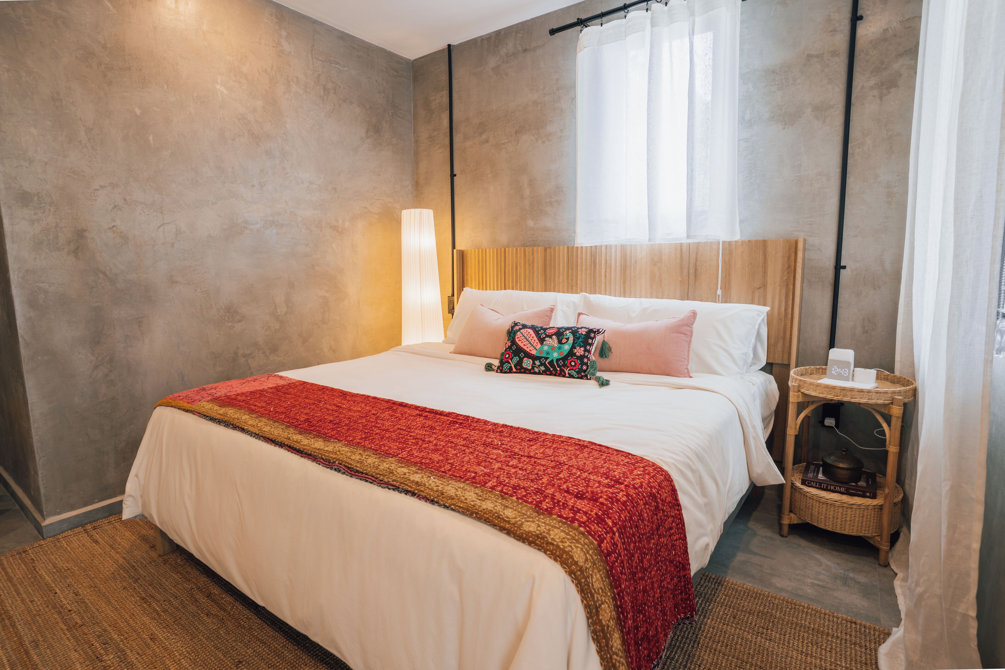 A modern bedroom with a large bed, a wooden headboard, a bedside table with a lamp, and a wicker side table.